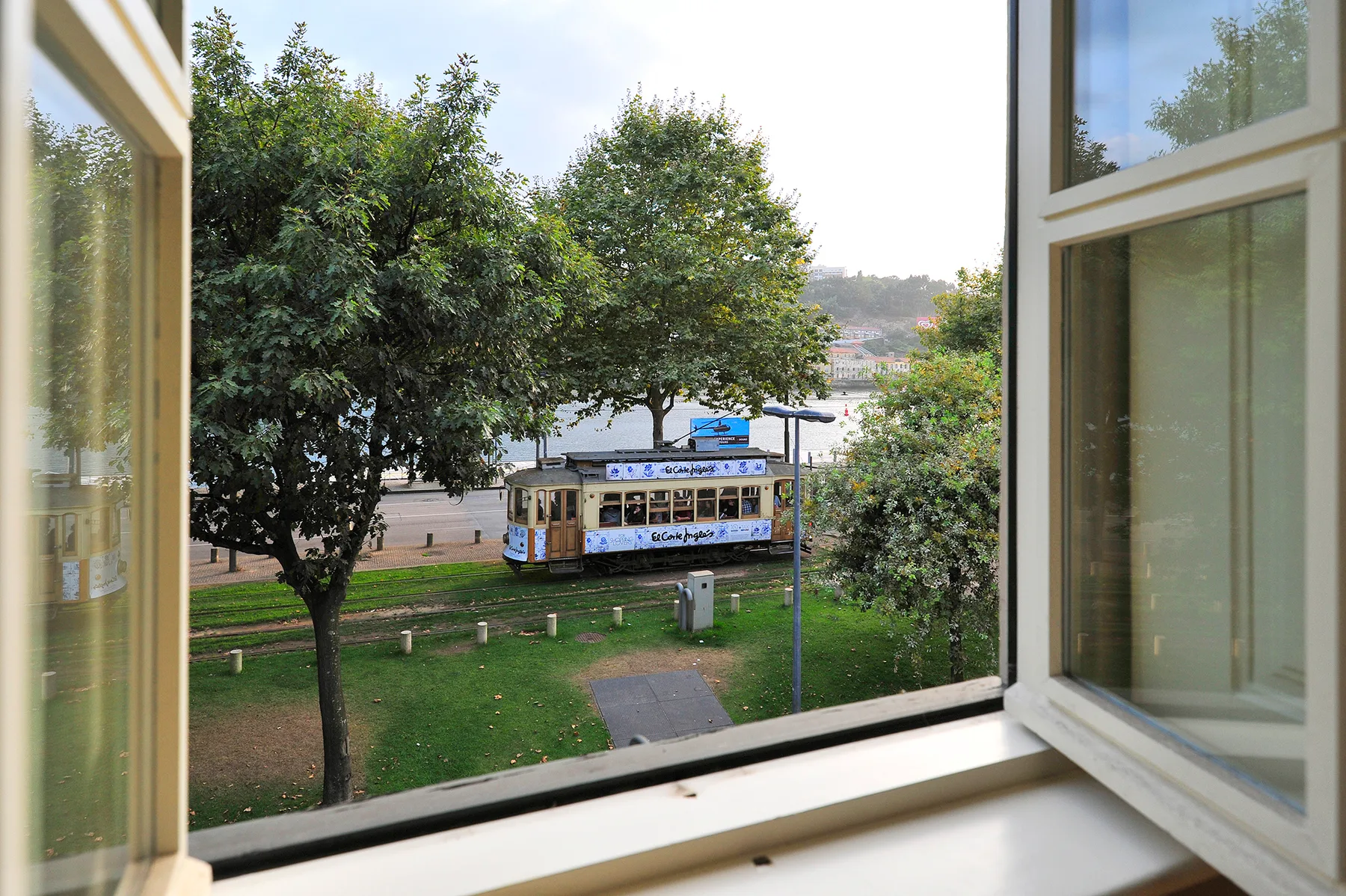 Vista desde una ventana abierta hacia un tranvía junto a un parque verde.