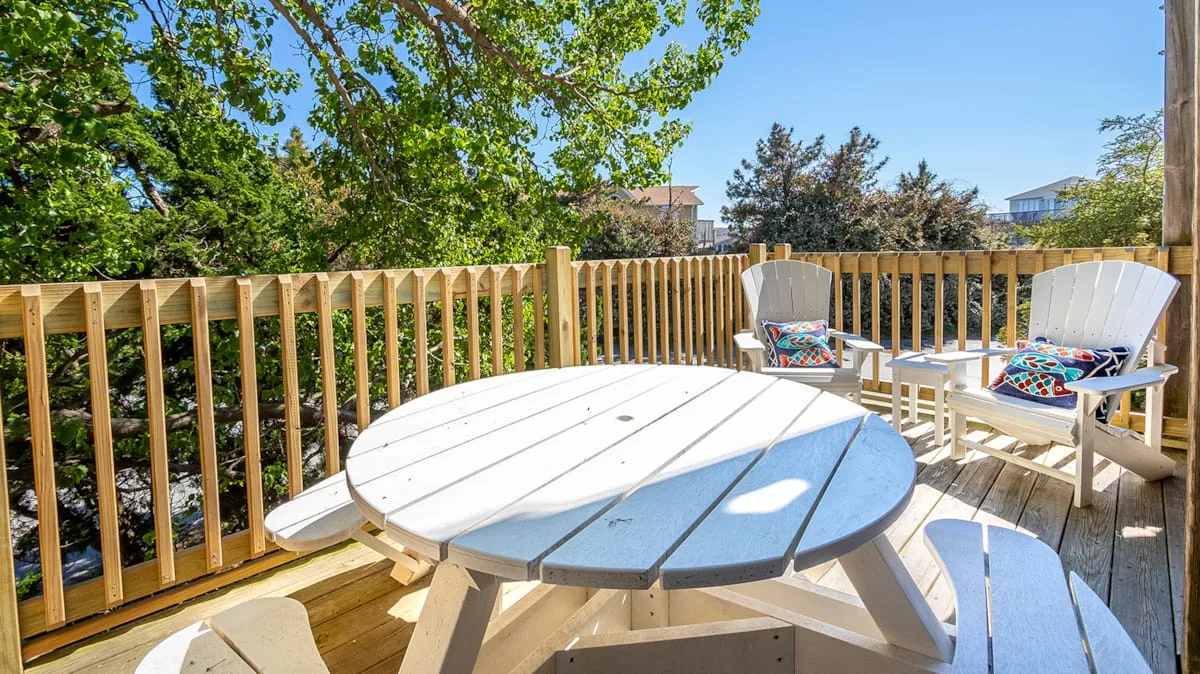 A sunny wooden deck has a round table and outdoor seating beneath leafy trees.