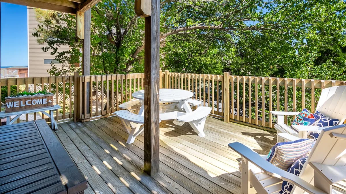 A wooden deck has picnic seating and lounge chairs beside leafy trees.