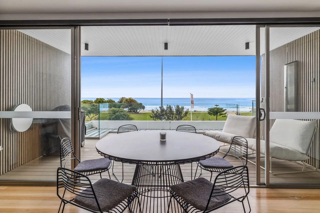 A dining area opens onto a balcony with outdoor seating and a coastal view.