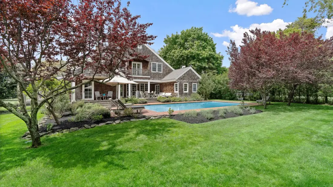 Una casa y una piscina exterior están junto a un amplio césped verde y árboles frondosos.