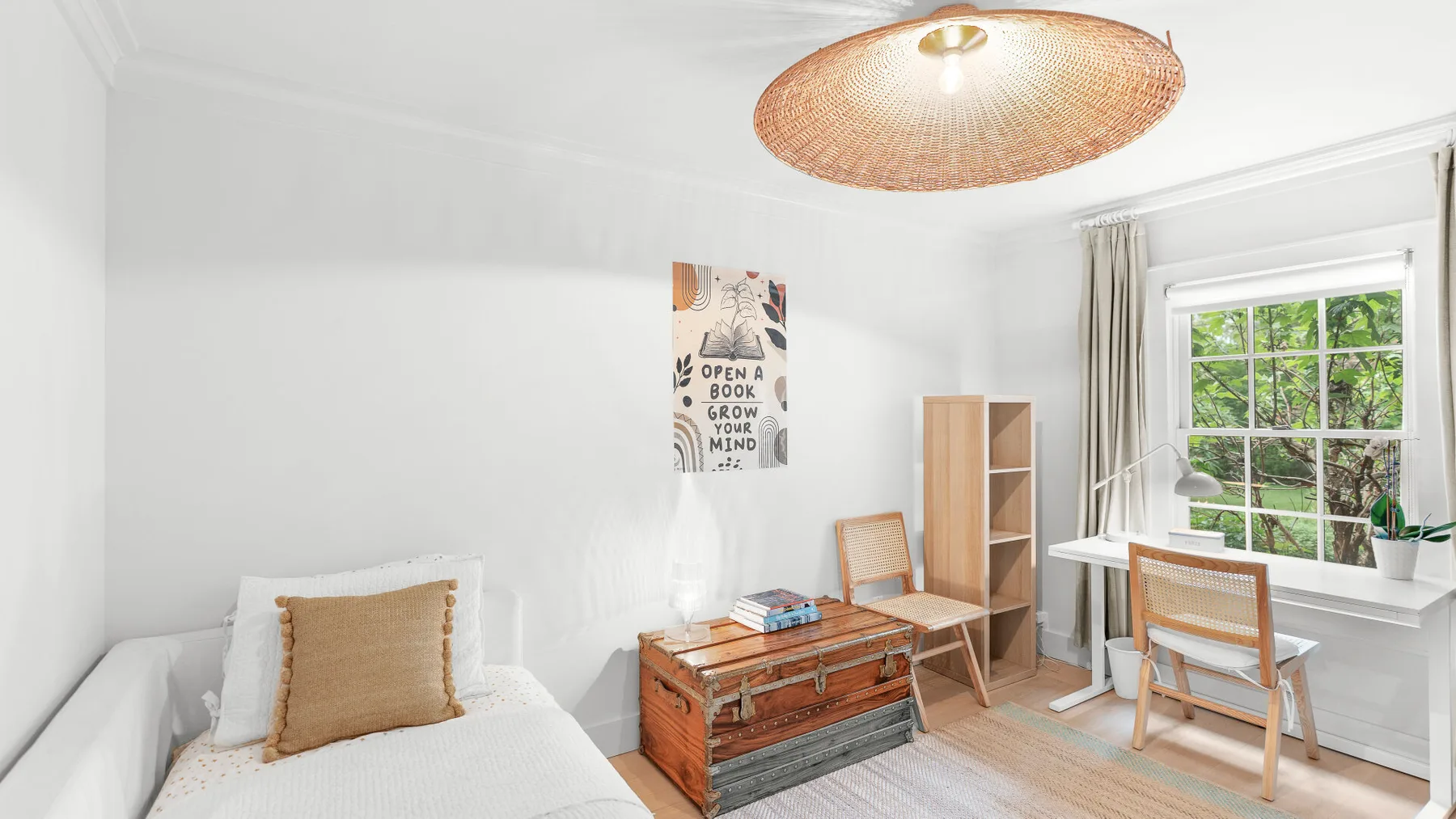 Un dormitorio luminoso con una cama, un baúl de madera y un escritorio junto a la ventana.