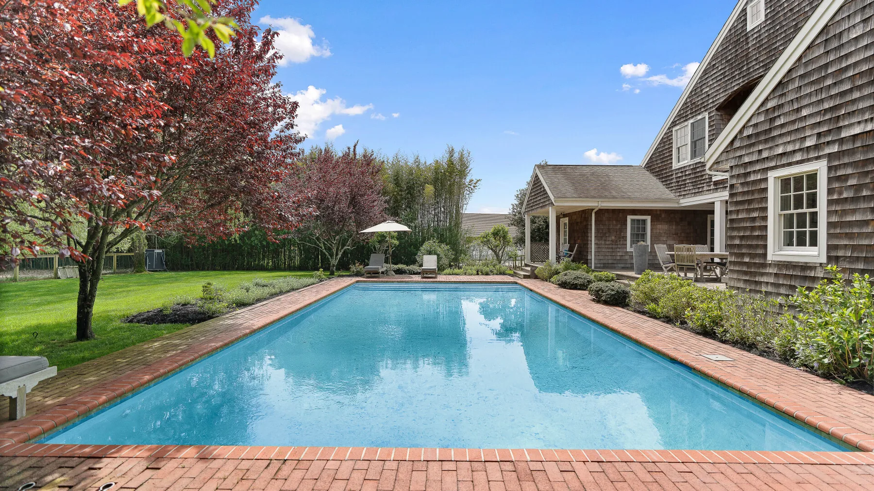 Una piscina junto a una casa grande con revestimiento de tejas y un jardín cuidado.