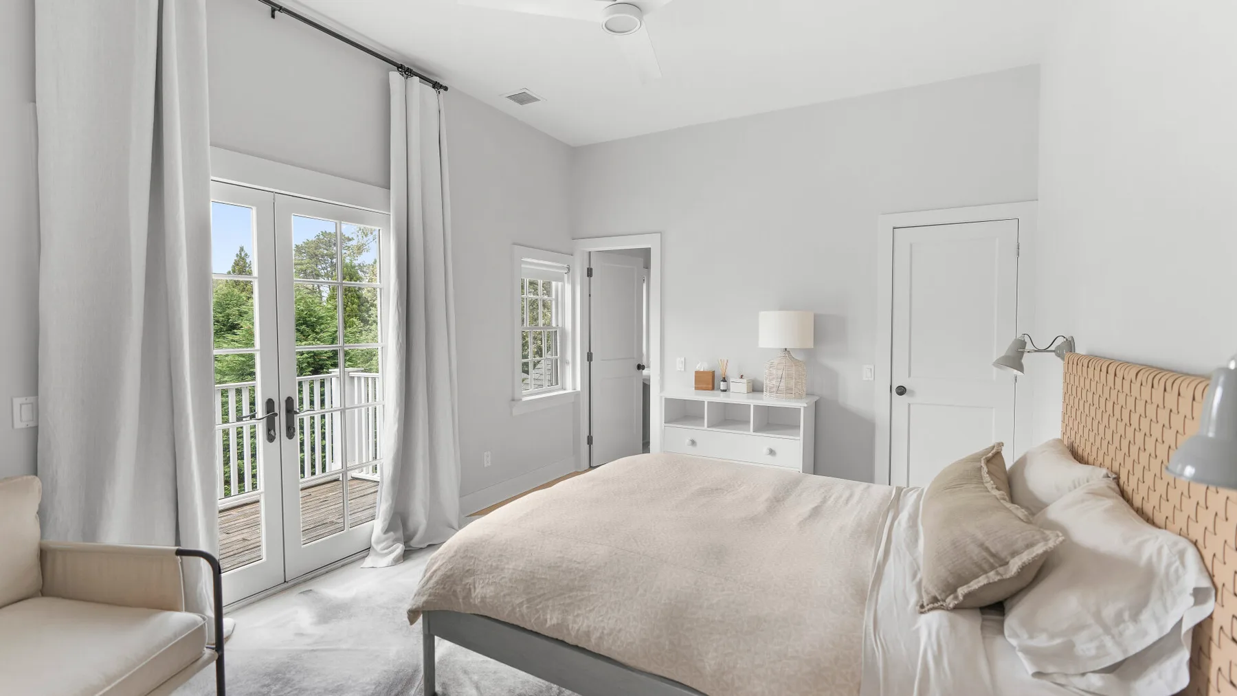 Un dormitorio luminoso con una cama, paredes claras y puertas de vidrio que dan a un balcón.