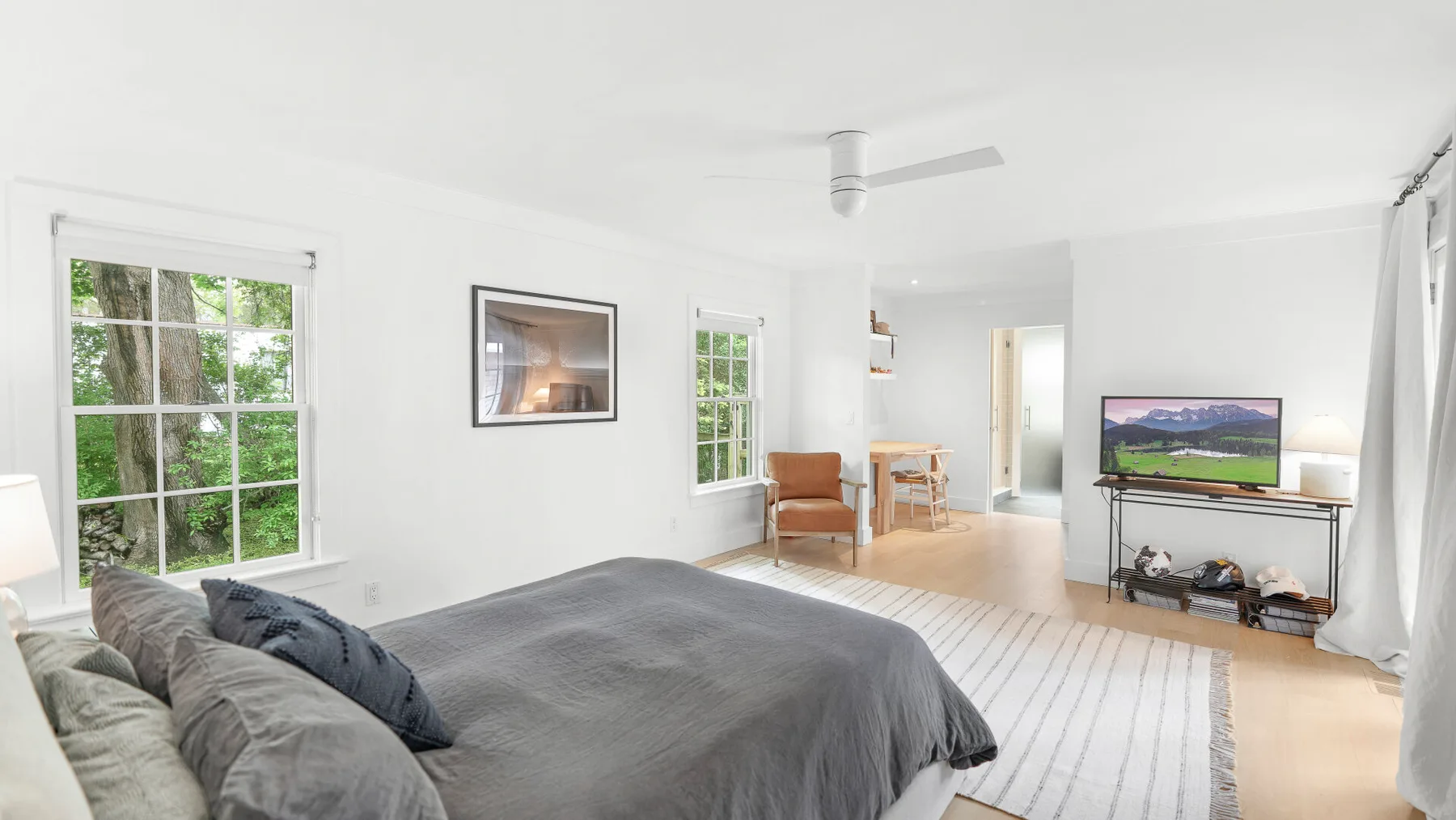 Una sala luminosa con un sofá gris, televisor y ventanas con vistas a la vegetación.