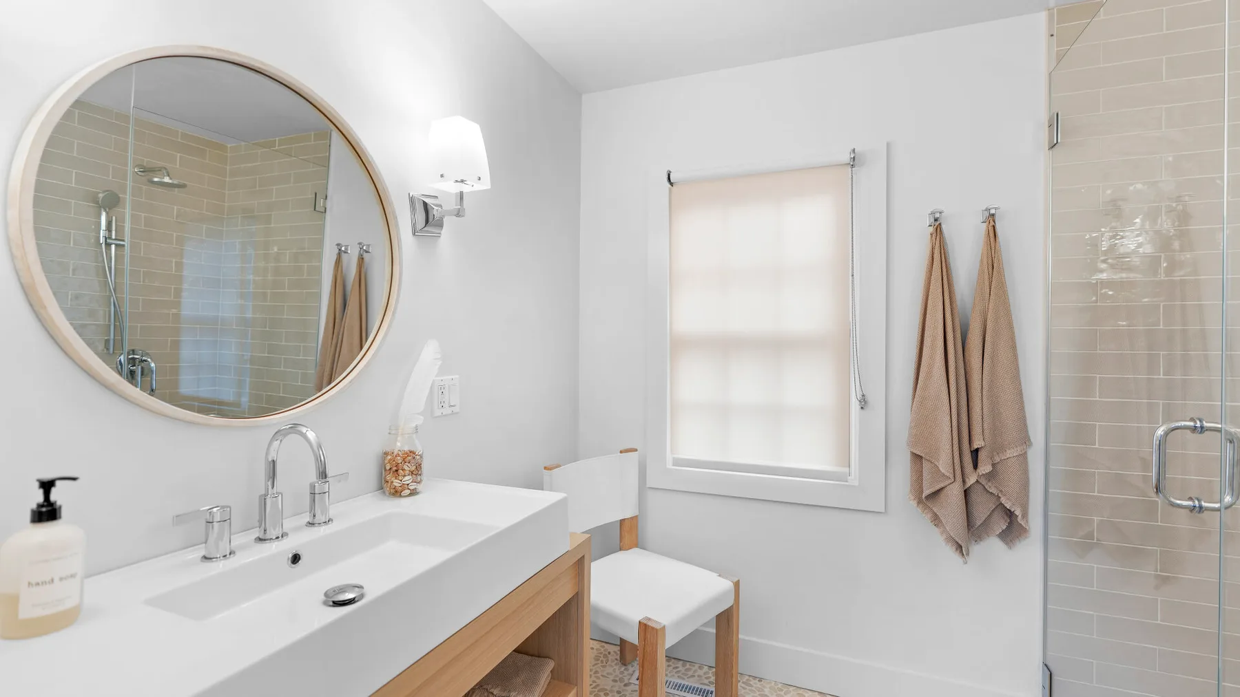 Un lavabo de baño luminoso está bajo un espejo redondo, junto a una ventana y una ducha.