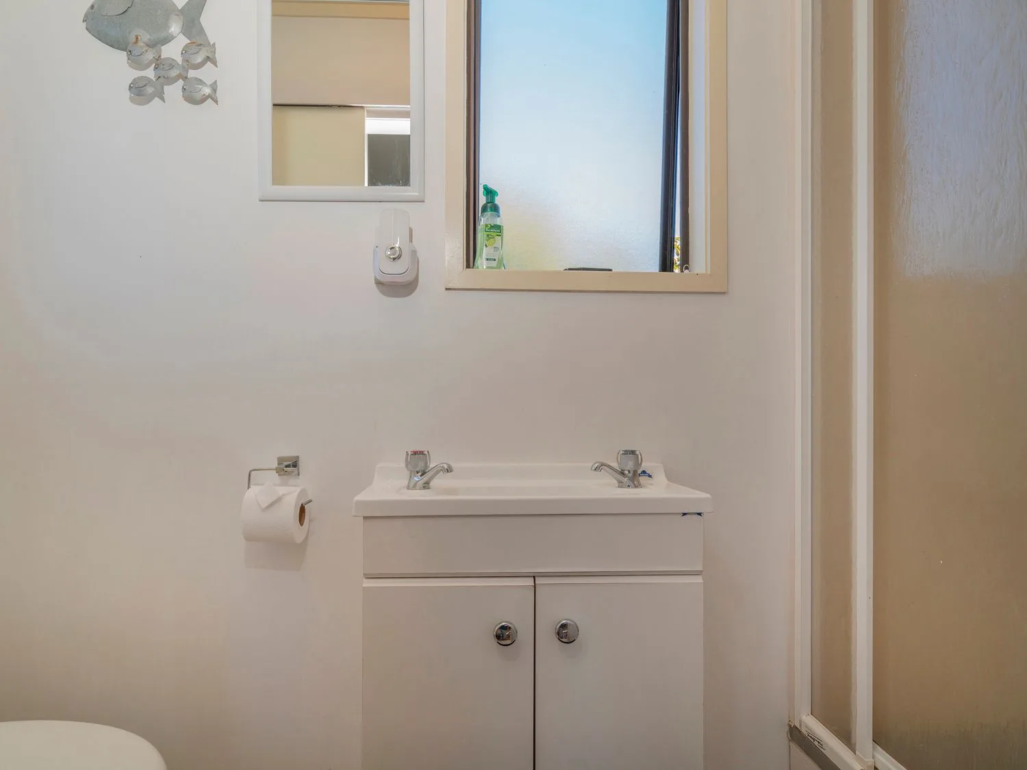 A bathroom sink with two taps mirror and window above a toilet paper holder with a roll and a shower door to the side at Slice of Pauanui Pauanui