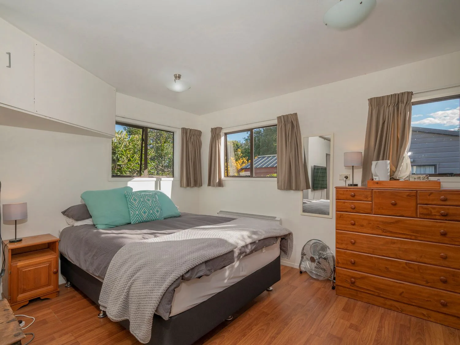 A bedroom with a bed and pillows beside a wooden nightstand and a wooden dresser under windows with curtains at Slice of Pauanui - Pauanui Holiday Home in Paunui
