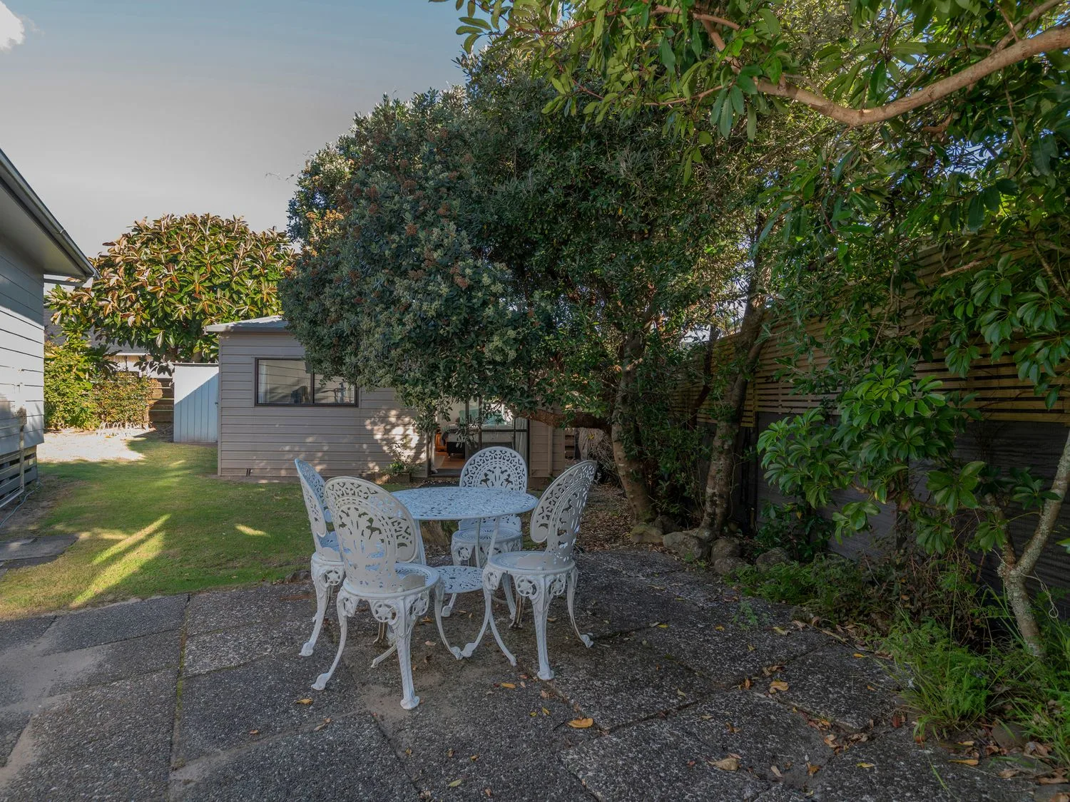 A garden patio with outdoor seating sits beneath leafy trees beside small buildings.