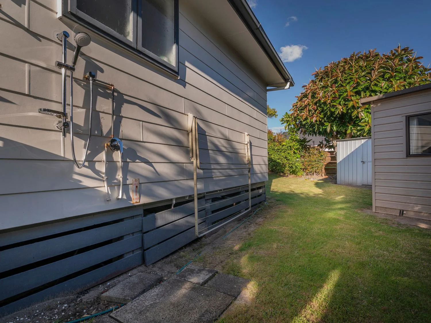An outdoor shower is mounted on the side of a house beside a grassy yard.