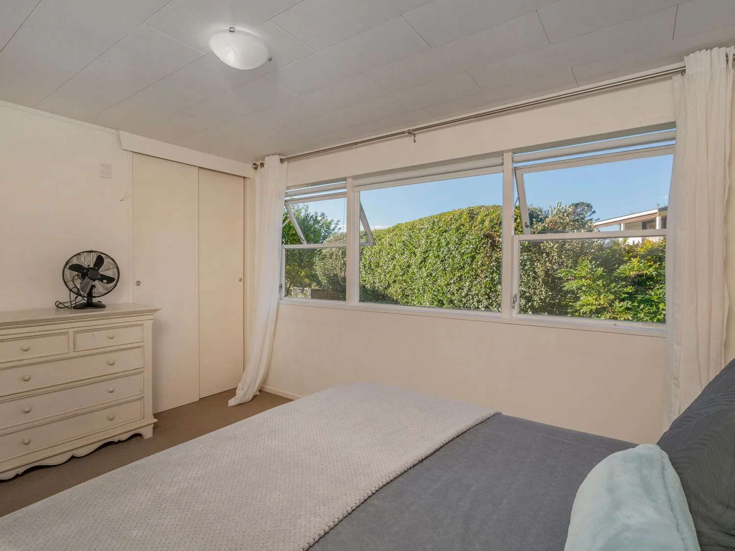 A bedroom with a bed a dresser with a fan and windows showing greenery outside at Slice of Pauanui - Pauanui Holiday Home in Paunui