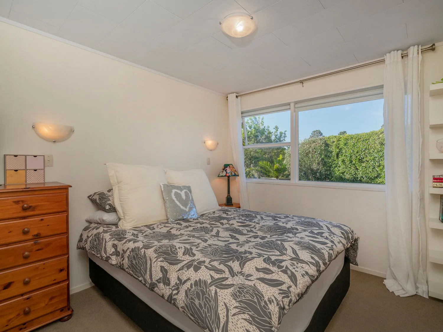 A bedroom with a bed a wooden chest of drawers and large window with white curtains at Slice of Pauanui - Pauanui Holiday Home Paunui