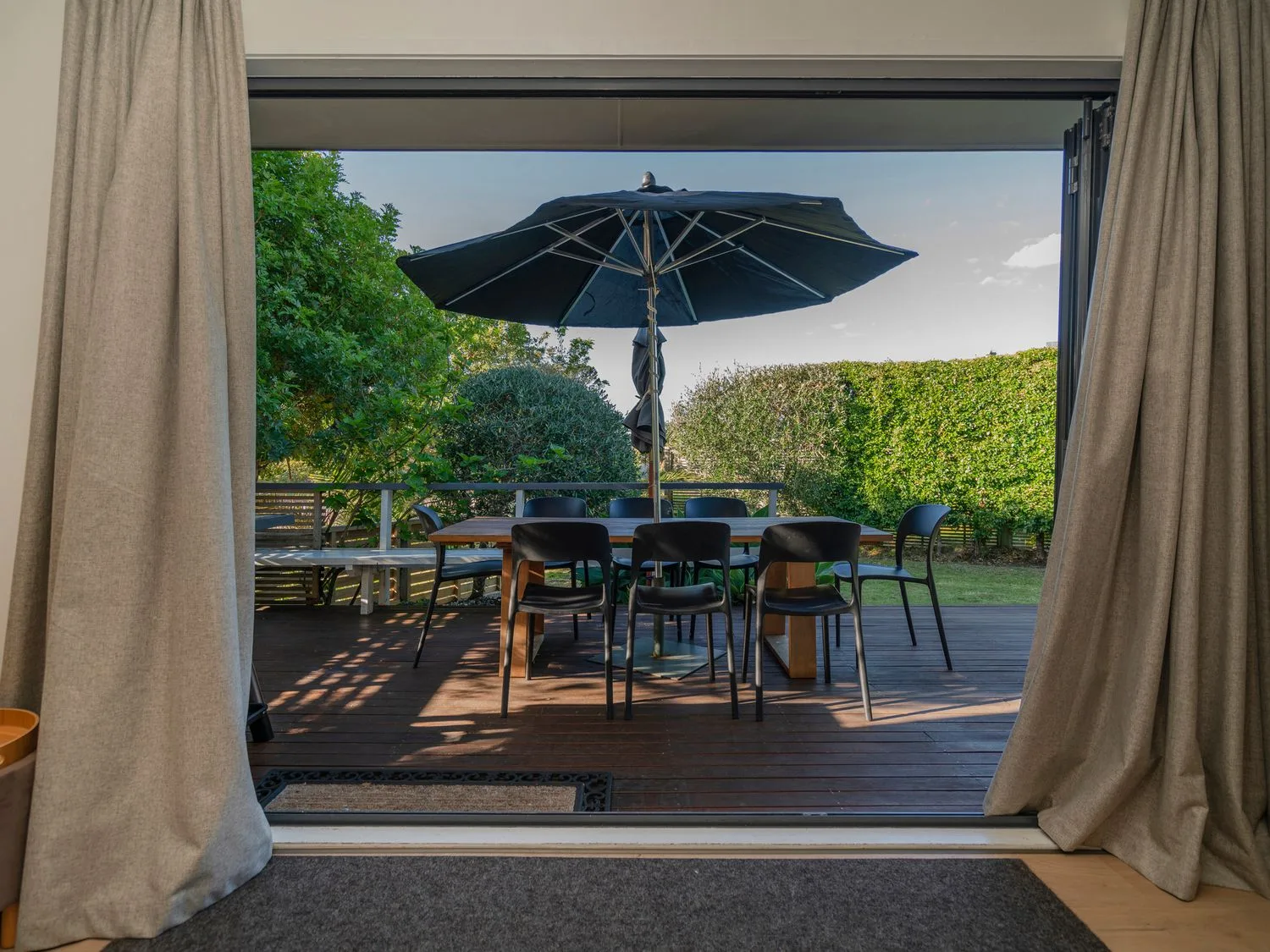 An outdoor dining area with a table, chairs and a large umbrella on a wooden deck surrounded by greenery at Slice of Pauanui - Pauanui Holiday Home in Paunui