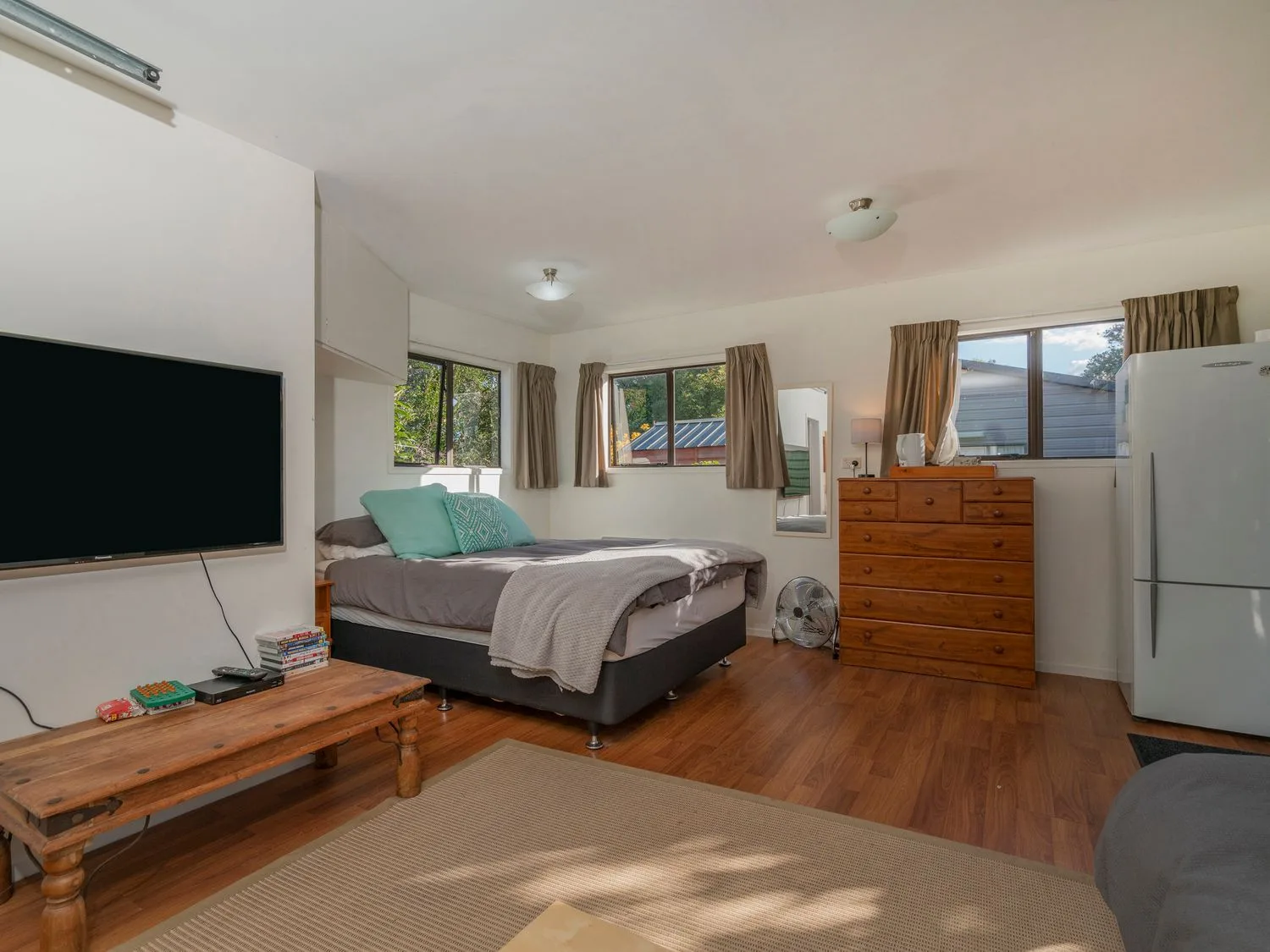 A bedroom with a bed a wooden dresser a refrigerator a wall-mounted TV and windows with curtains at Slice of Pauanui - Pauanui Holiday Home in Paunui