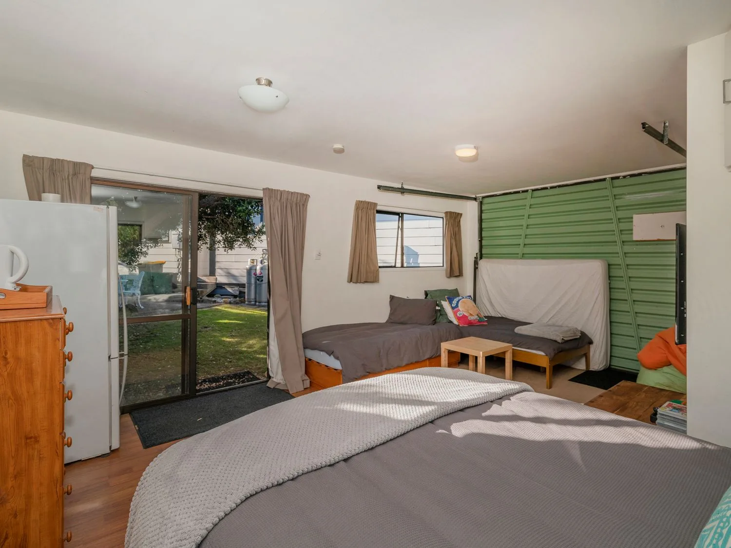 A bedroom with beds, a refrigerator, and a sliding glass door opening to a garden.