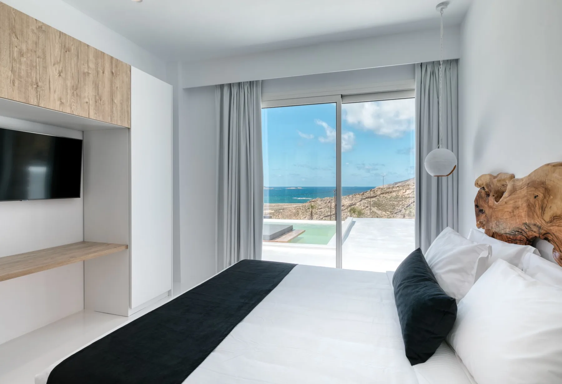 A bedroom with a bed beside a large window overlooking the sea.