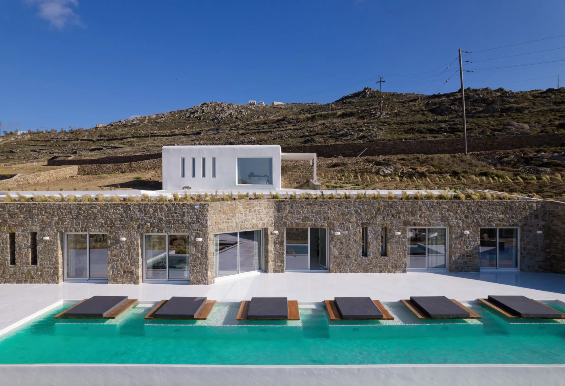 A modern stone villa opens onto a turquoise swimming pool beneath a rocky hillside.