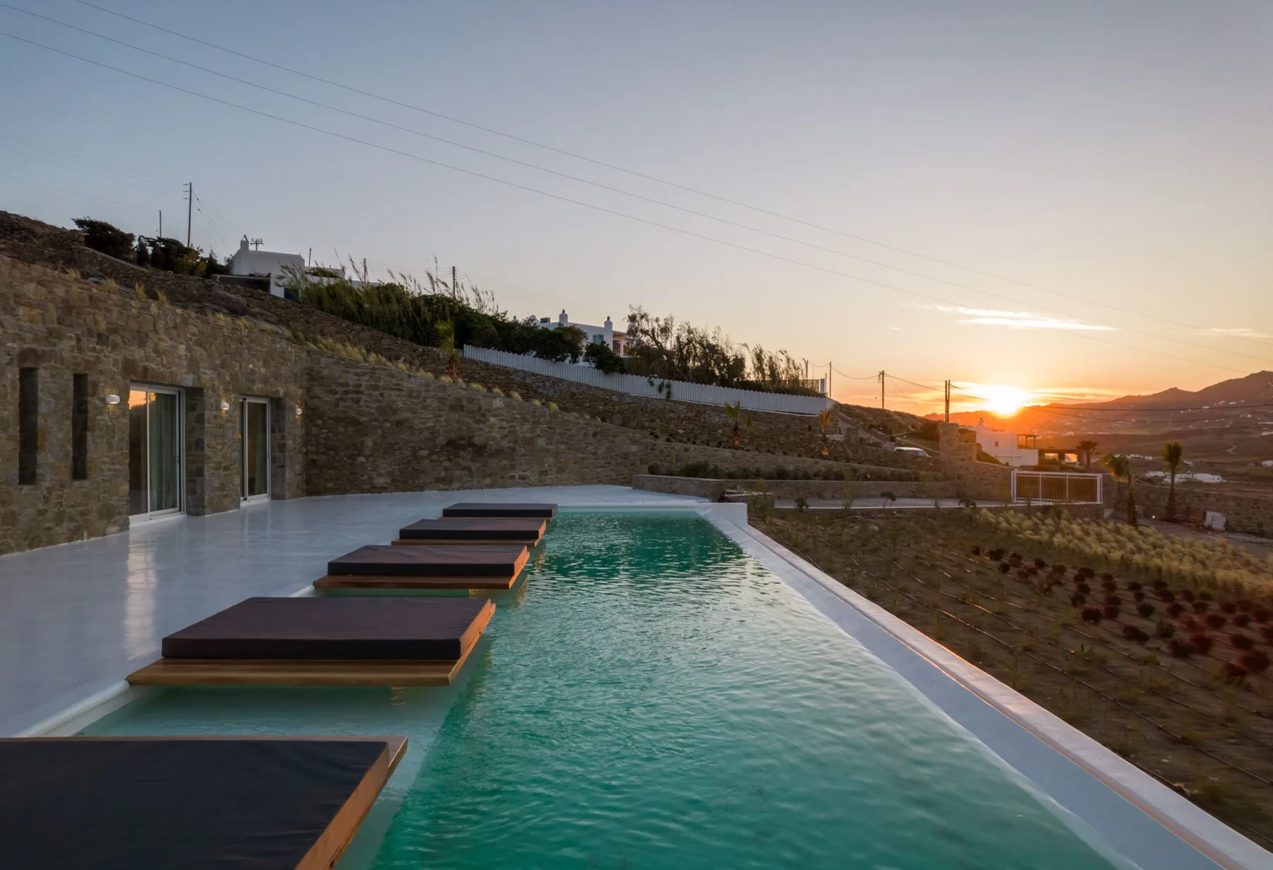 An outdoor swimming pool sits beside a stone wall at sunset.