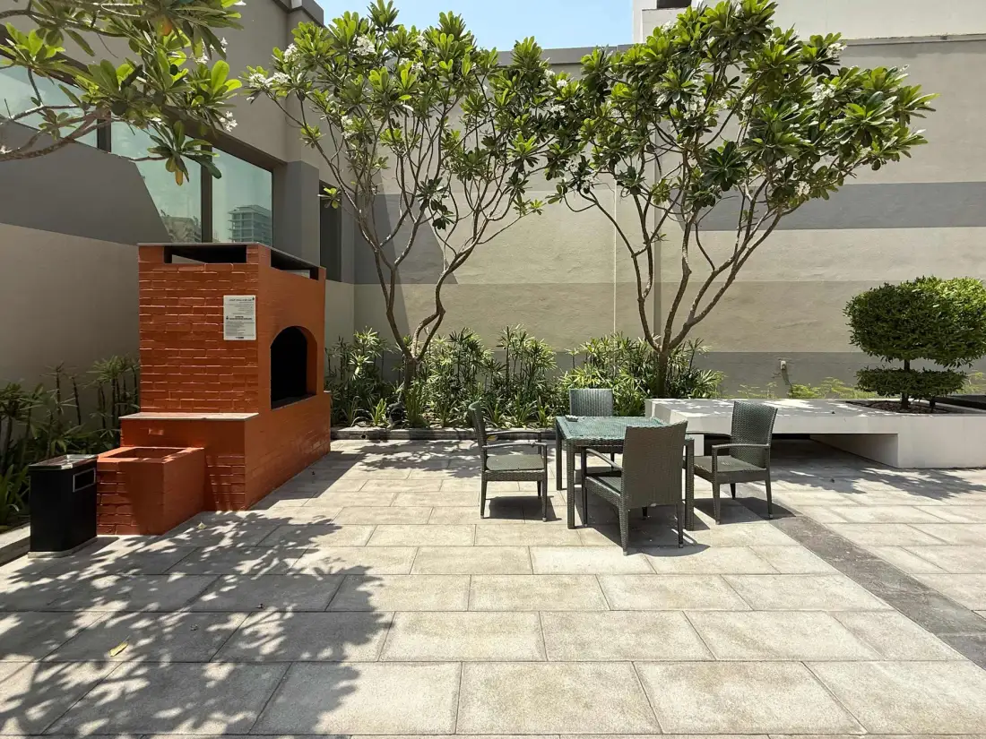 Patio exterior pavimentado con mesa, sillas, árboles y una parrilla de ladrillo.
