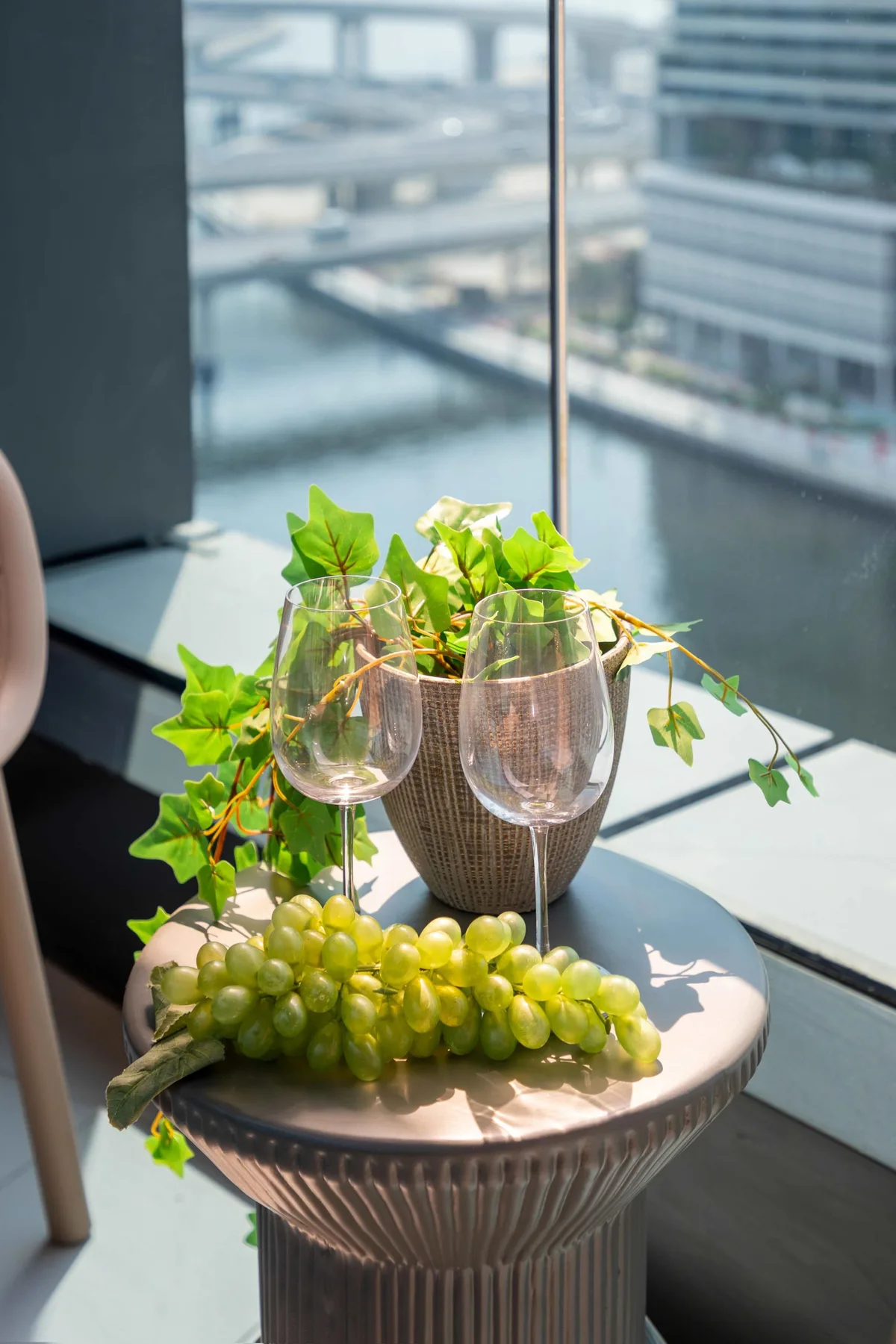 Una escena junto a una ventana muestra una planta en maceta, dos copas y uvas sobre una mesa pequeña.