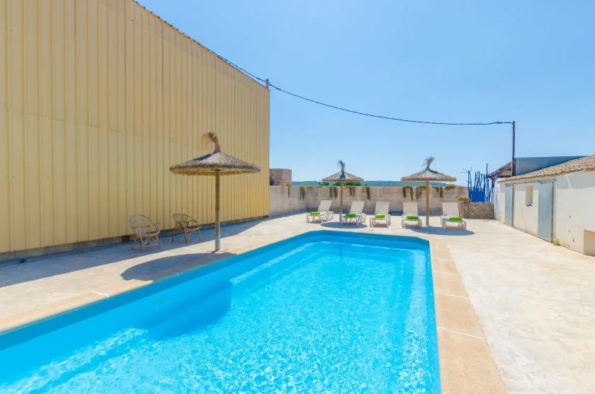 Una piscina exterior luminosa está junto a una zona pavimentada con asientos a la sombra.