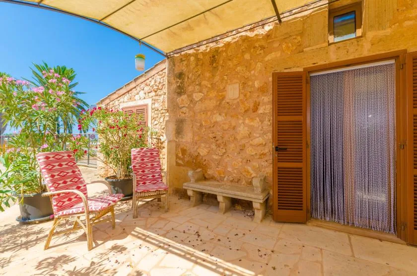 Un patio soleado de piedra tiene asientos a rayas rojas, plantas en macetas y una entrada sombreada.