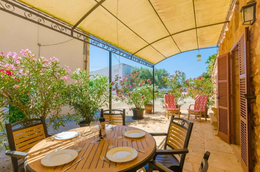 Comedor exterior a la sombra con mesas de madera, asientos y plantas en flor.