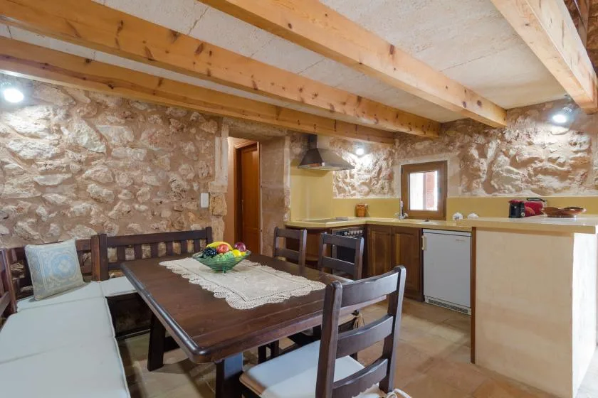 Un comedor rústico comunica con una cocina, con una mesa de madera bajo vigas expuestas.