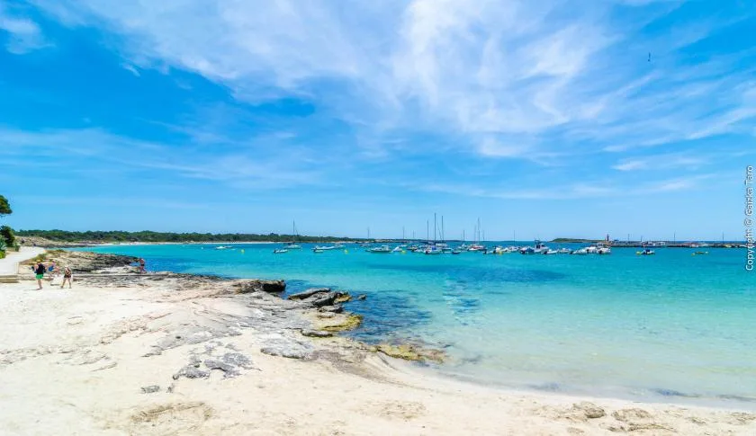 Una playa de arena bordea aguas turquesas y claras bajo un cielo azul.