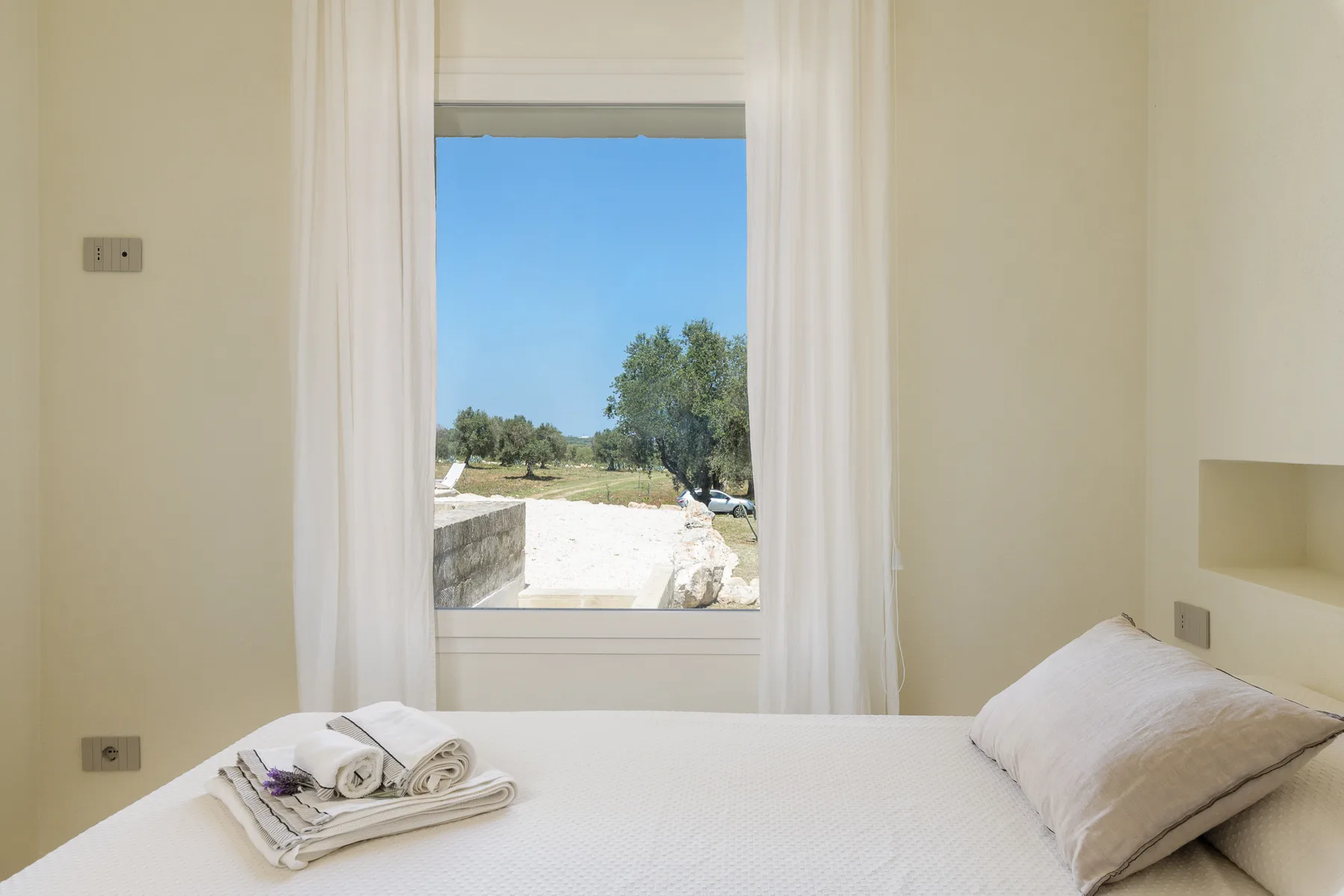 A bedroom with a bed beside a window overlooking trees and open ground.