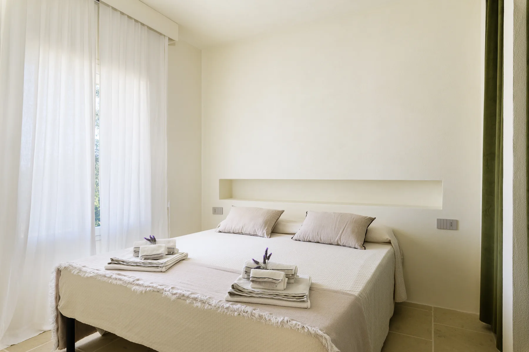 A bright bedroom with a neatly made bed beside sheer curtains.