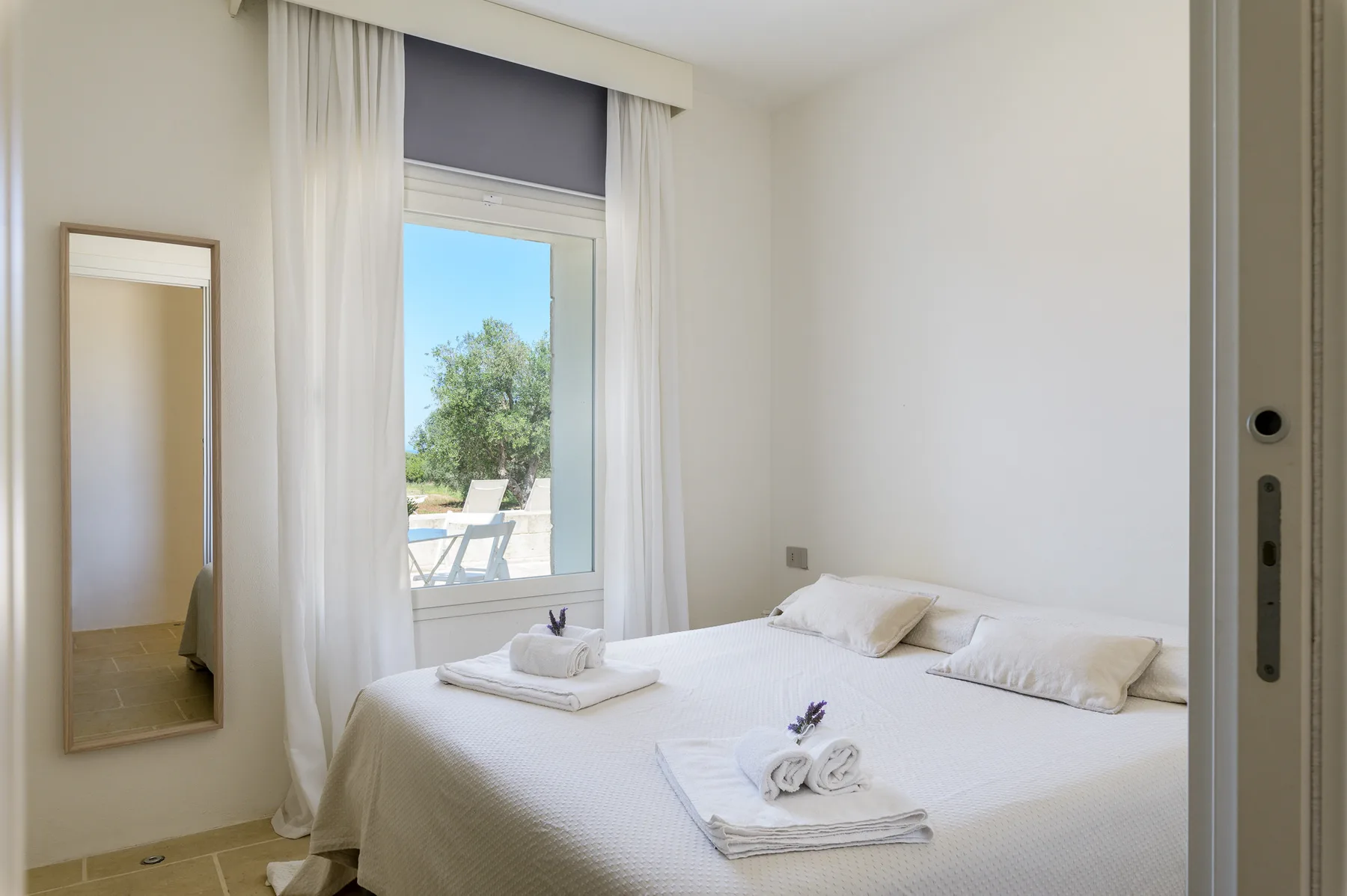 A bright bedroom with a neatly made bed beside a curtained window.