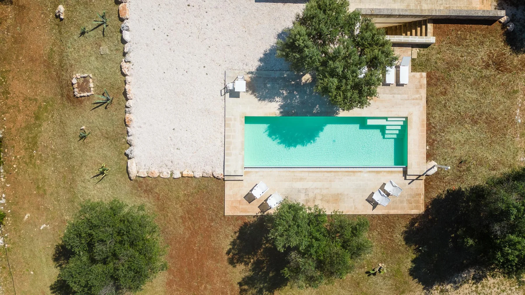 An outdoor swimming pool sits beside a pale paved area and surrounding trees.