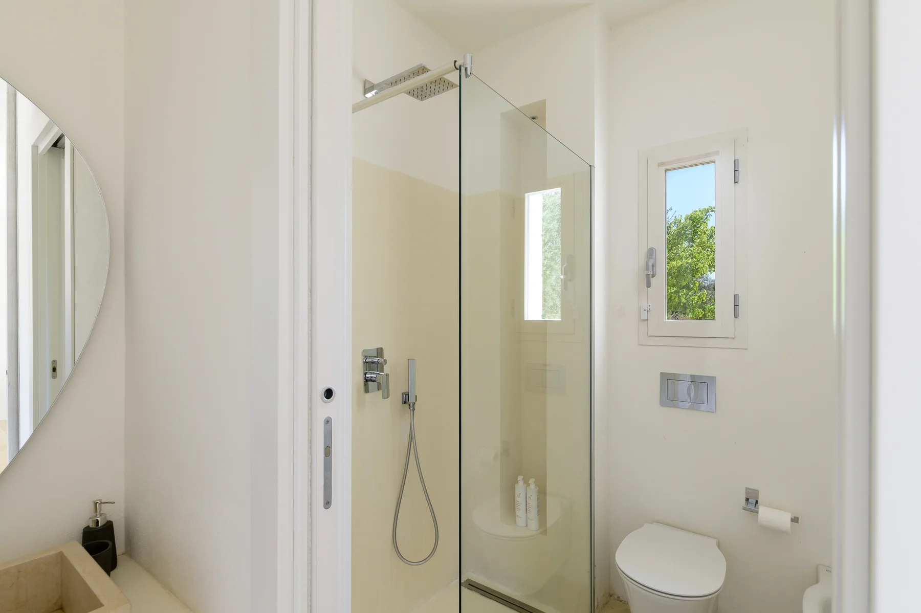 A bright bathroom with a glass shower, toilet, and small window.