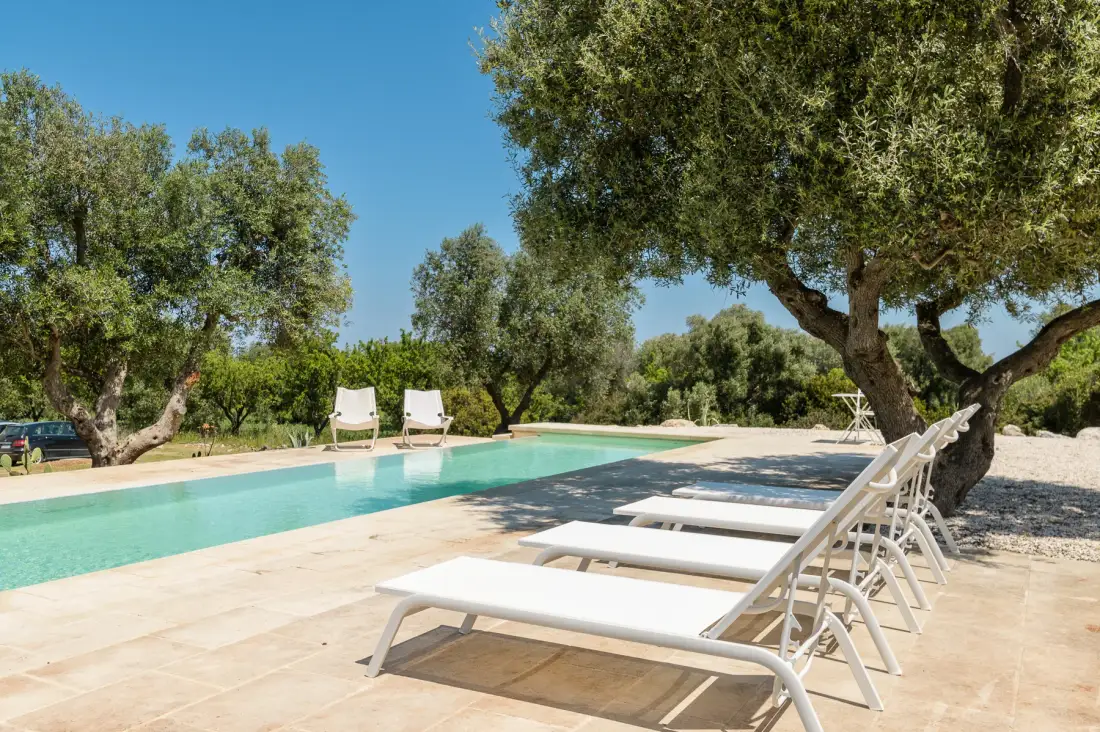 An outdoor swimming pool beside a paved deck with lounge seating and trees.
