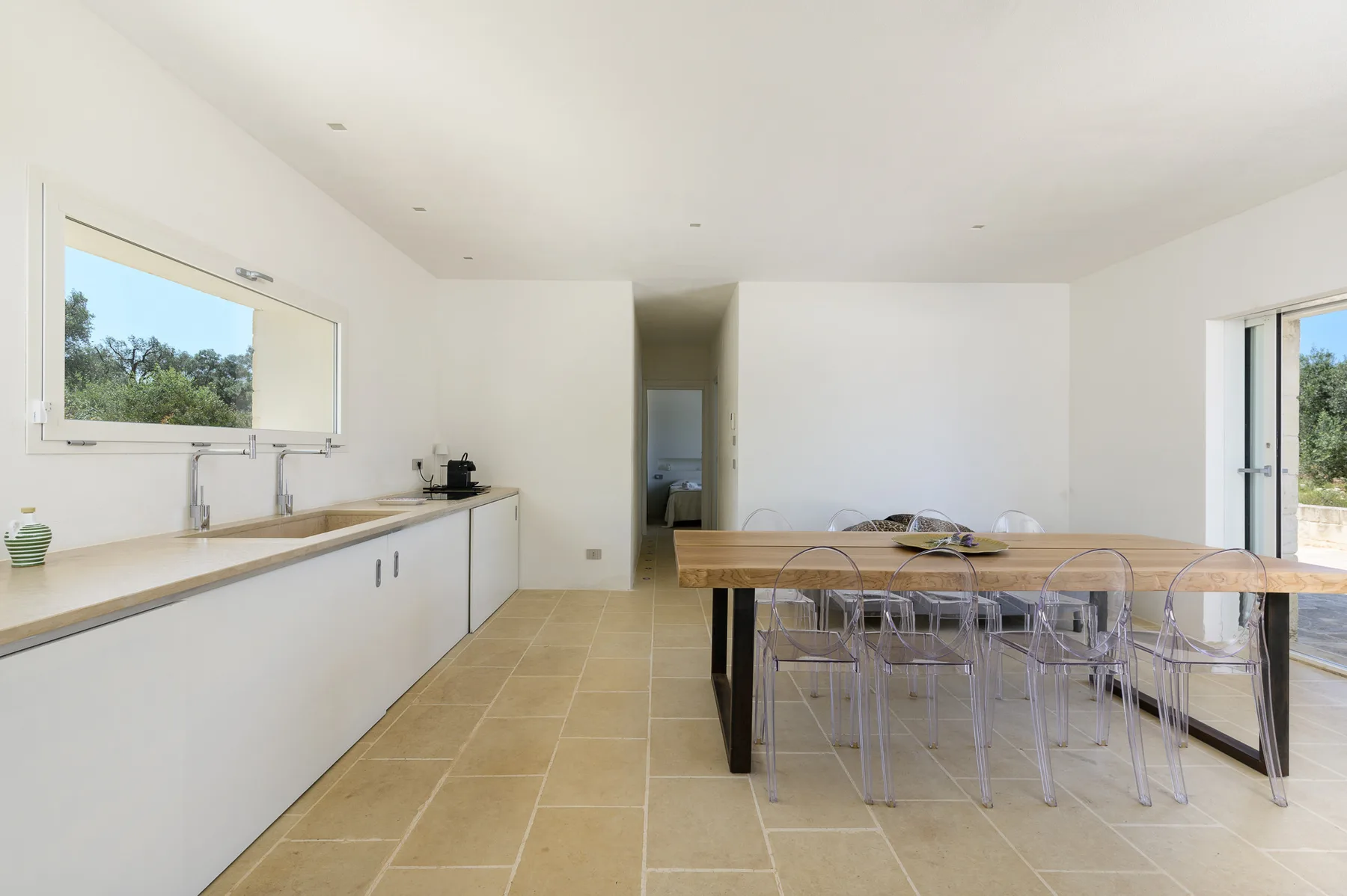 A bright dining area with a long table and clear chairs beside a kitchen counter.