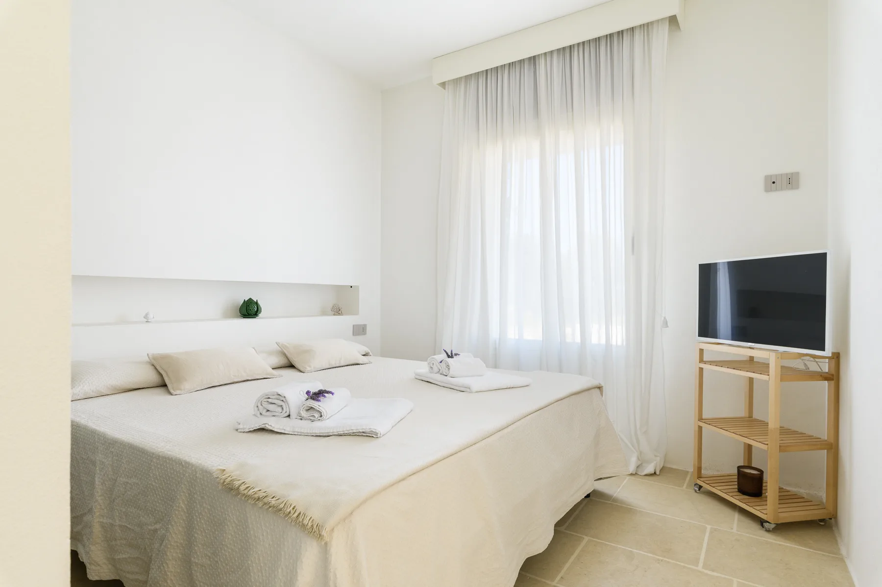 A bright bedroom with a neatly made bed and a television beside the window.