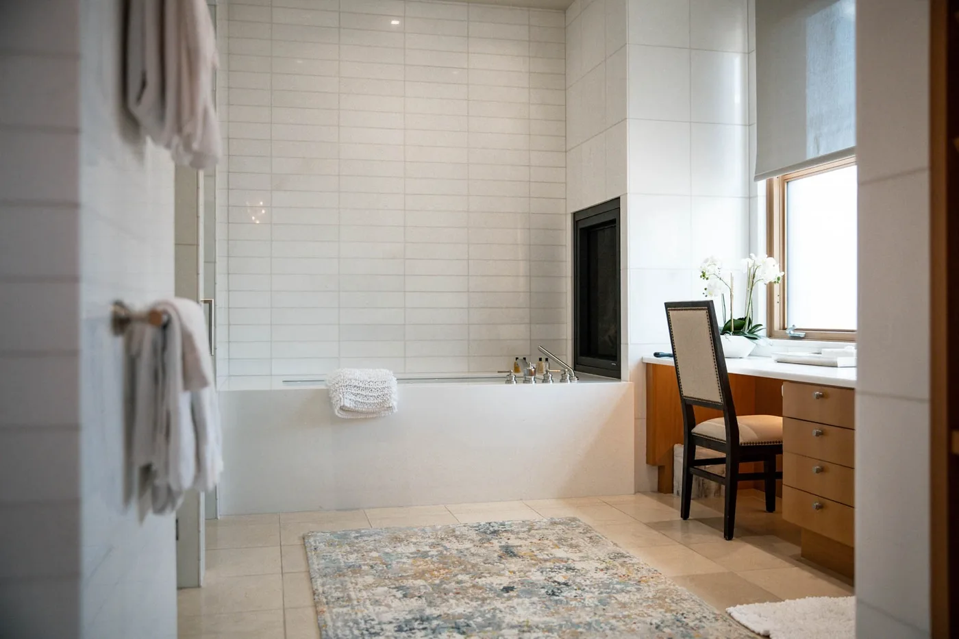 A tiled bathroom with a bathtub, vanity, and a chair beside the window.