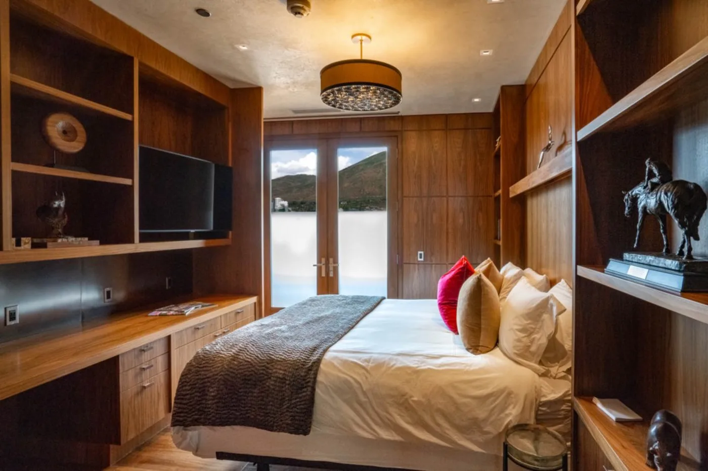A bedroom with a bed, built-in wooden shelving, and windows overlooking a hillside.