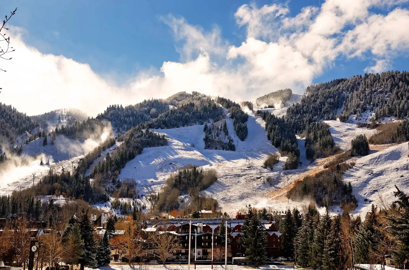 Snow-covered mountain slopes rise behind a wooded valley and buildings.