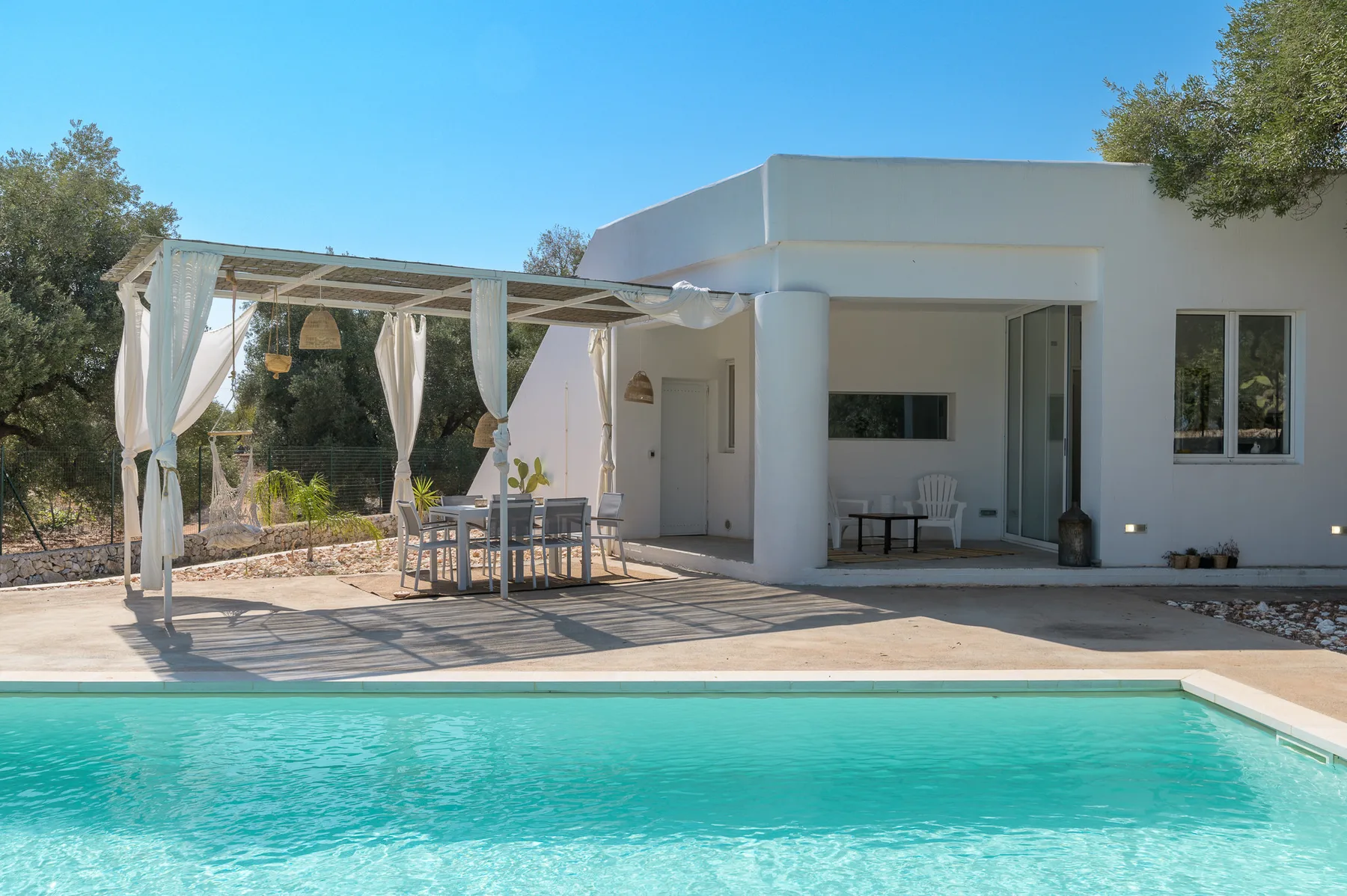 A swimming pool borders a modern white house with a shaded outdoor seating area.