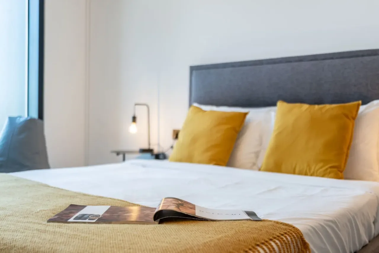 Un dormitorio con una cama tendida, almohadas amarillas y una manta doblada.