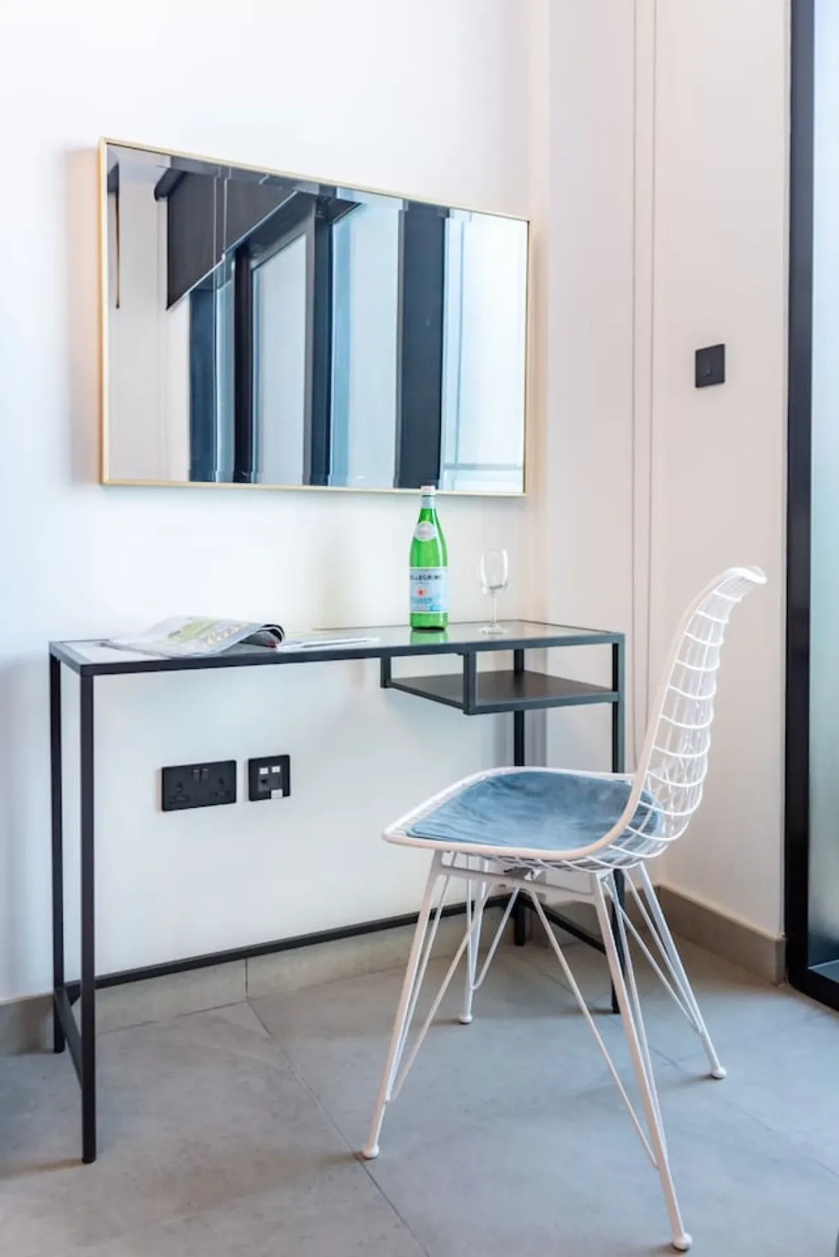 A small desk area with a clear chair, wall mirror, and bottle on the desk.