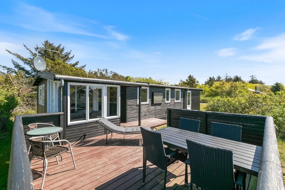 Una terraza de madera junto a una cabaña moderna tiene asientos y una mesa de comedor.