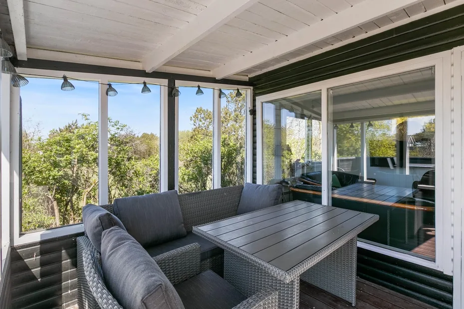 Un patio cubierto con asientos tejidos y una mesa junto a grandes ventanas.