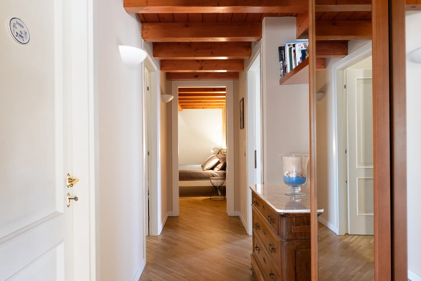 A hallway with wooden ceiling beams, a wooden floor, and a bedroom visible at the end.