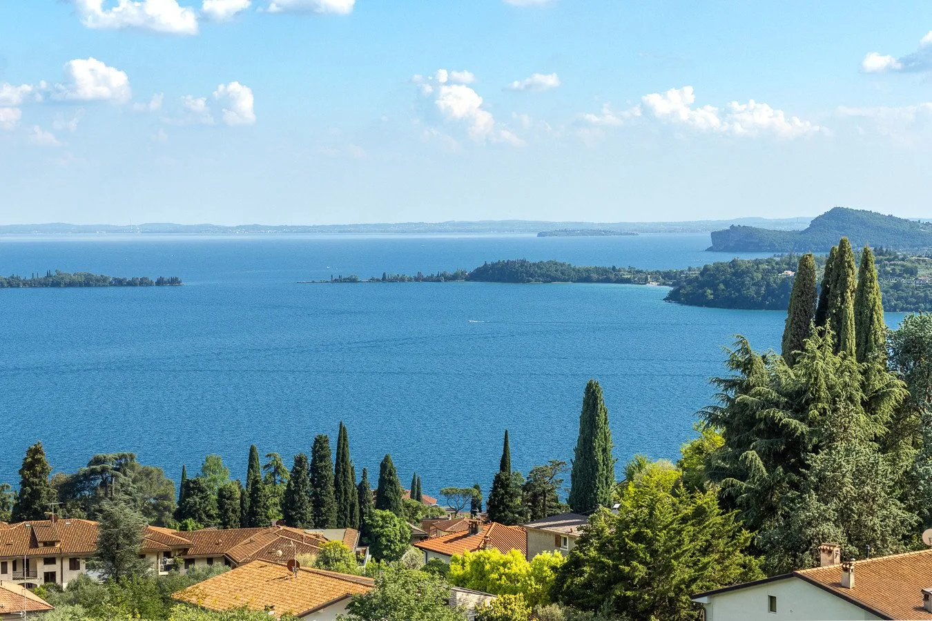 A broad blue lake stretches beyond trees and red-roofed buildings under a partly cloudy sky.