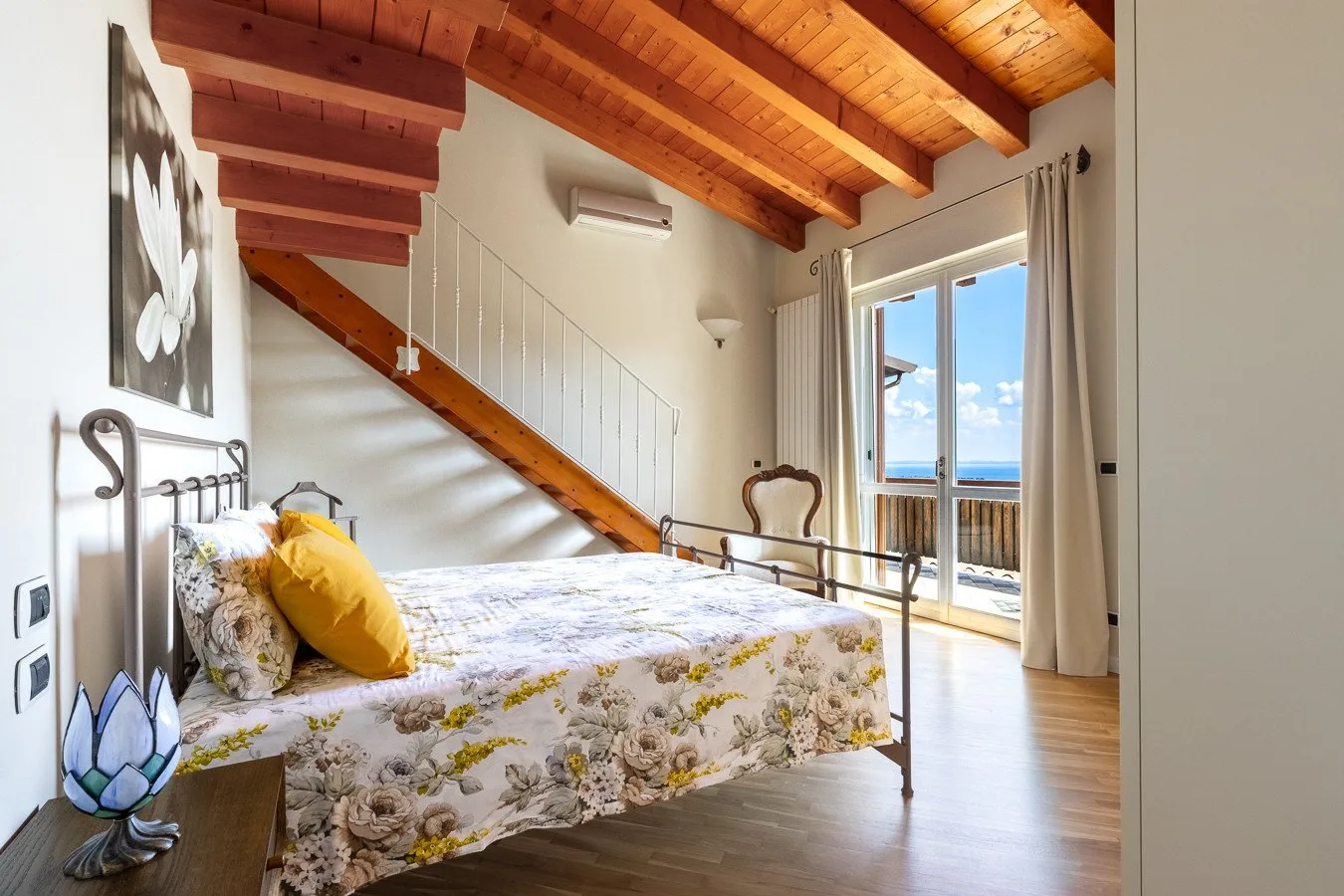 A bedroom with a floral bedspread and a staircase beneath a wooden ceiling.