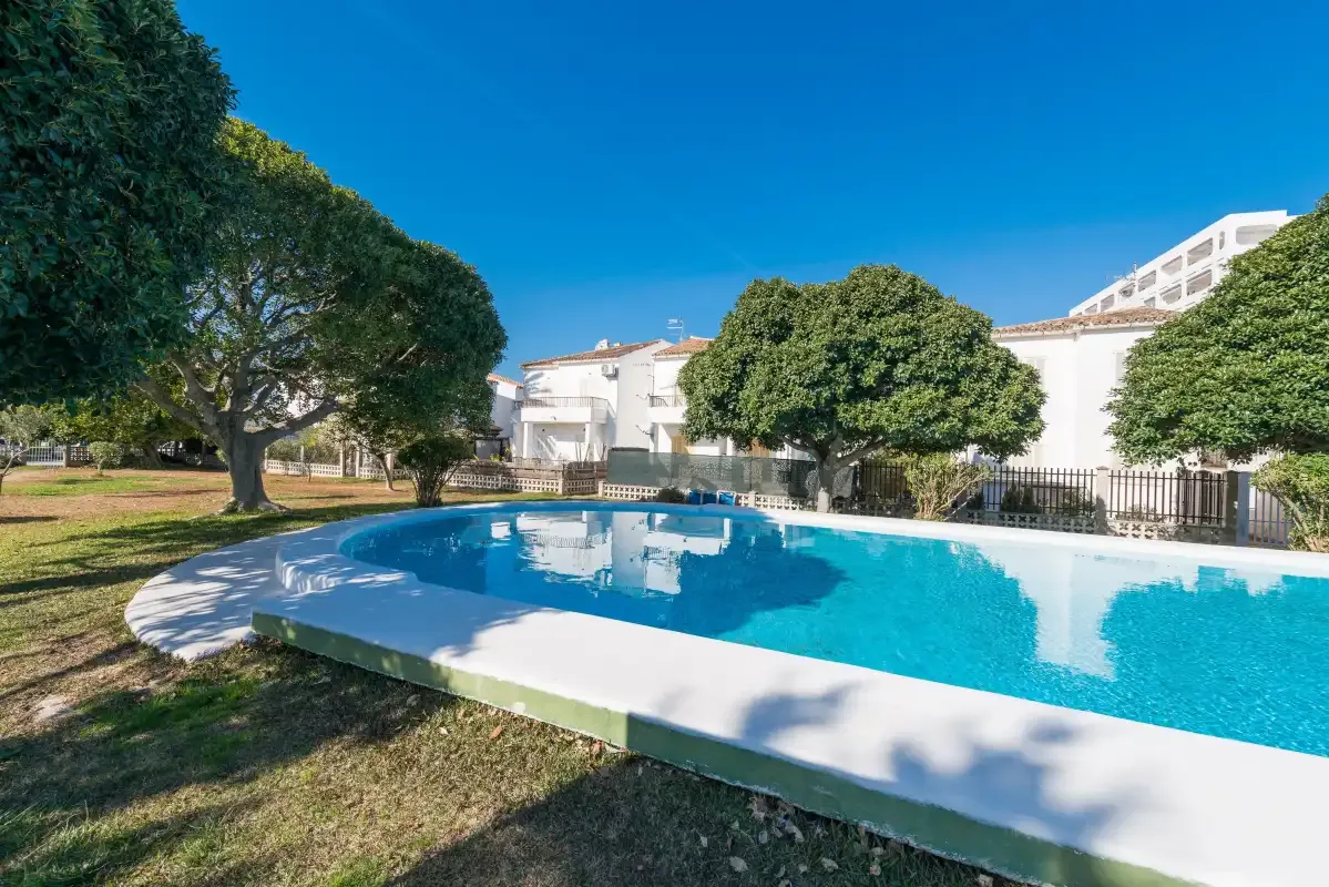 An outdoor swimming pool sits beside trees and white buildings under a clear sky.