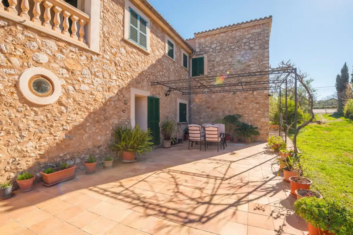 Un patio de piedra soleado tiene asientos al aire libre y plantas en macetas junto a la casa.